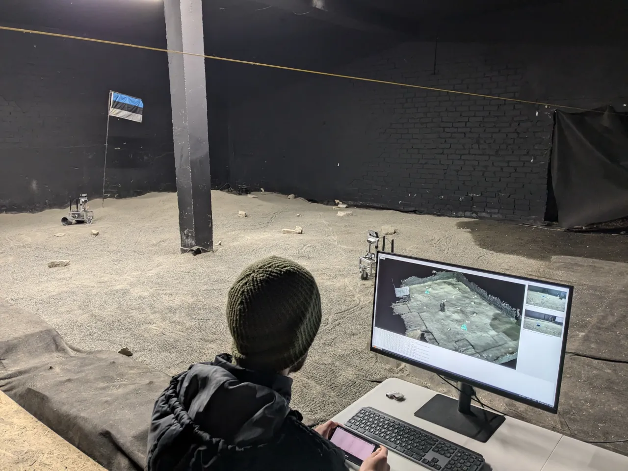 An operator at a monitor showing a live 3D map, with a rover on the sand behind