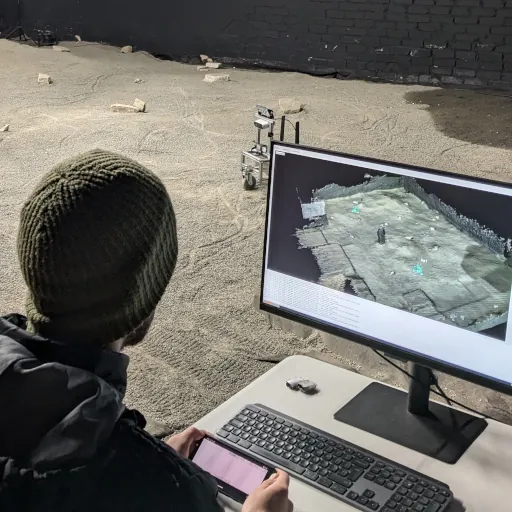 An operator at a monitor showing a live 3D map, with a rover on the sand behind